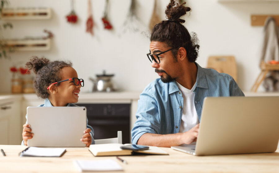 Young african american father works remotely on laptop while his little son using digital tablet